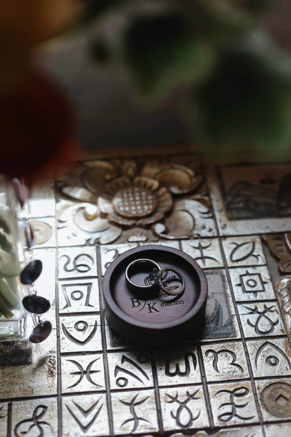 A wooden wedding ring holder with engraved initials and two rings on an ornate Spanish tile table.
