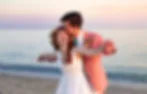 A couple celebrates at the beach; the woman in a white dress laughs joyfully as the man in a pink jacket kisses her cheek at sunset.