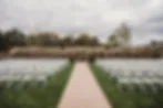 Outdoor wedding setup with empty white chairs facing an archway surrounded by tall grasses. Overcast sky adds a calm mood.