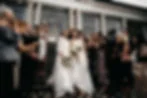 Two women in white dresses holding bouquets, joyfully descending stairs surrounded by applauding guests, with petals in the air. Elegant building backdrop.