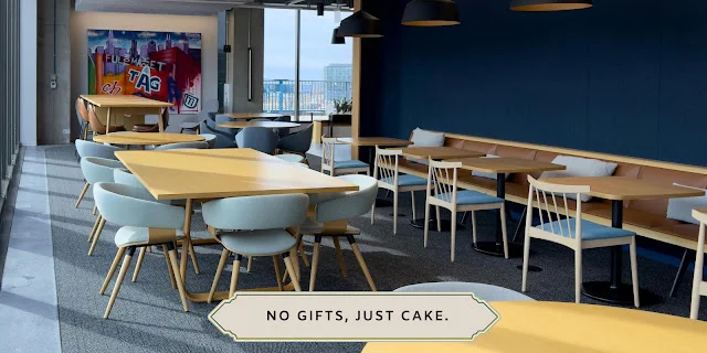 No Gifts, Just Cake: Workplace Wedding Celebration Etiquette Modern corporate office lounge with light wood tables and blue chairs, featuring a minimalist text overlay that reads No Gifts, Just Cake.