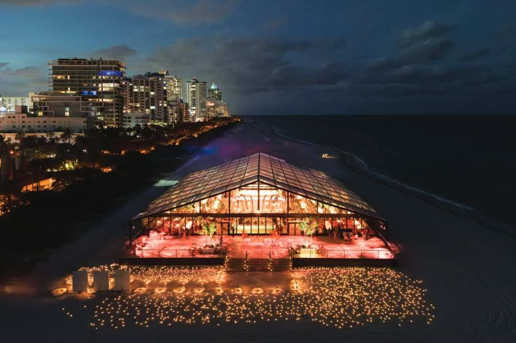 Beachside wedding venue with illuminated glass pavilion and city skyline at dusk.