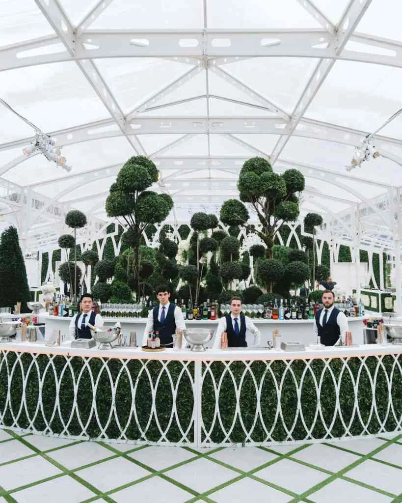Elegant bar setup with five bartenders in a glass conservatory with topiary trees.