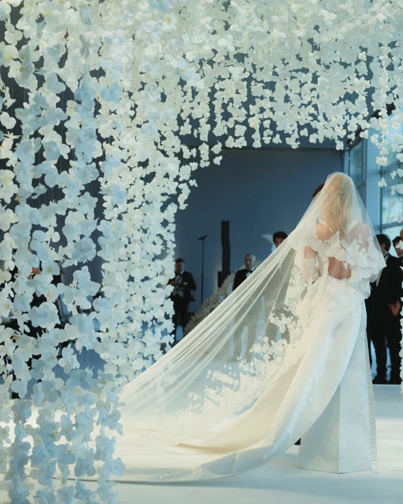 Bride in a white wedding gown with long veil under floral arch at wedding reception.