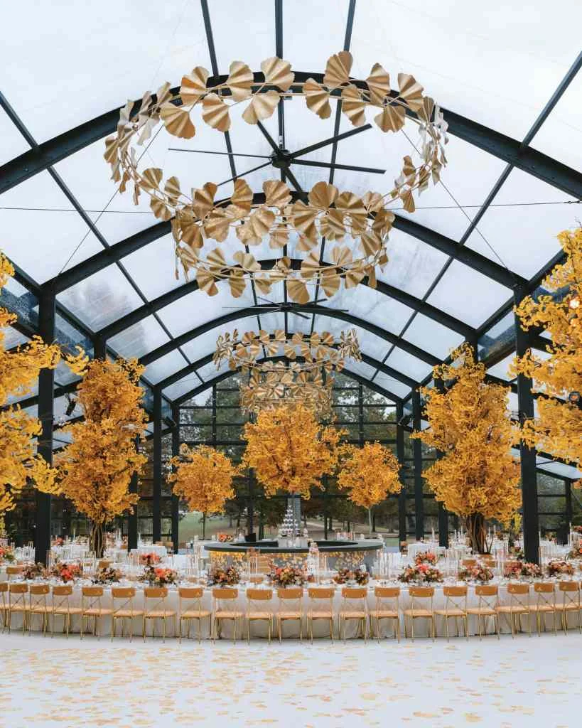 Elegant wedding reception with autumn trees and floral decor in glass conservatory.